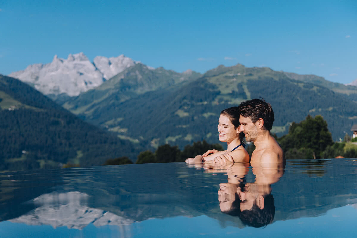 Ein Mann und eine Frau entspannen im Infinity-Pool. Im Hintergrund erstrecken sich grüne Hügel und schneebedeckte Berge unter klarem, blauem Himmel.