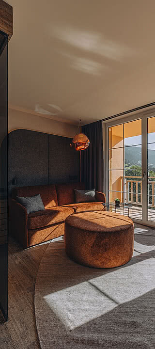 CSP_08092024_Hotel_Fernblick_116.dng Wohnzimmer mit Sauna, orangefarbenem Sofa, rundem Hocker und Schreibtisch. Große Fenster bieten Bergblick und führen zu einem Balkon. Dezente Beleuchtung und Holzdachakzente.