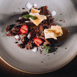 Dessert bestehend aus Schokoladenkekskrümeln, Erdbeerscheiben, Sahnetupfern und Minzblättern liegt auf einem großen, grauen Teller. Lichteinfall von rechts erzeugt Schatten auf dem Tisch.