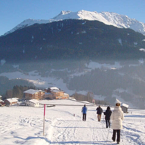 Menschen spazieren auf schneebedecktem Weg, umgeben von alpinen Holzhäusern und hohen, schneebedeckten Bergen unter klarem, blauem Himmel.