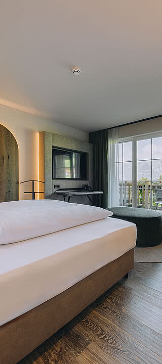 CSP_08092024_Hotel_Fernblick_156.dng Ein modernes Hotelzimmer mit großem Bett, daneben eine gemütliche Sitzecke. Große Fenster bieten Blick auf einen Balkon, stilvolle Beleuchtung und Holzböden ergänzen das Ambiente.