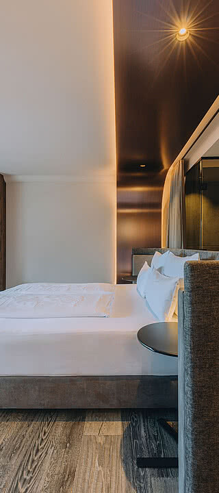 Bett mit weißer Bettwäsche steht in modernem Hotelzimmer aus Holz. Fernseher über Schreibtisch, Badezimmer mit offenen Regalen im Hintergrund. Dezente Beleuchtung erzeugt gemütliche Atmosphäre.