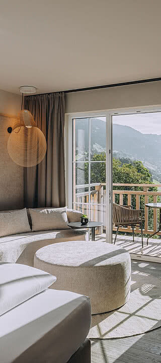 CSP_08092024_Hotel_Fernblick_096.dng Bett und Sofa stehen in einem hellen Zimmer mit offenem Balkon, der eine Aussicht auf Berge bietet. Ein runder Tisch und Schreibtisch ergänzen das moderne Interieur.