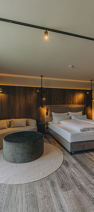 CSP_08092024_Hotel_Fernblick_208.dng Bett steht in modernem, elegantem Schlafzimmer mit Holzwänden, grauem Boden und großen Fenstern. Ein Sofa mit Ottomane und ein Schreibtisch ergänzen die Einrichtung.