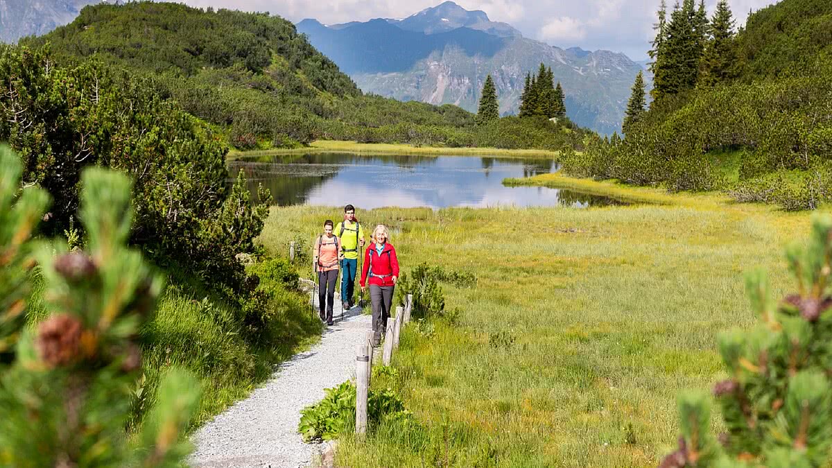 Drei Personen wandern auf einem Kiesweg durch eine grüne, leicht hügelige Landschaft mit einem See und Bergkulisse im Hintergrund unter bewölktem Himmel.