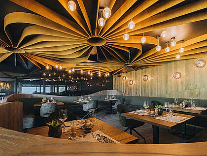 Ein stilvoll eingerichtetes Restaurant mit modernen Deckeninstallationen und schicker Beleuchtung. Tische sind elegant gedeckt, umgeben von gepolsterten Stühlen. Holzwände und gedämpftes Licht schaffen eine gemütliche Atmosphäre.