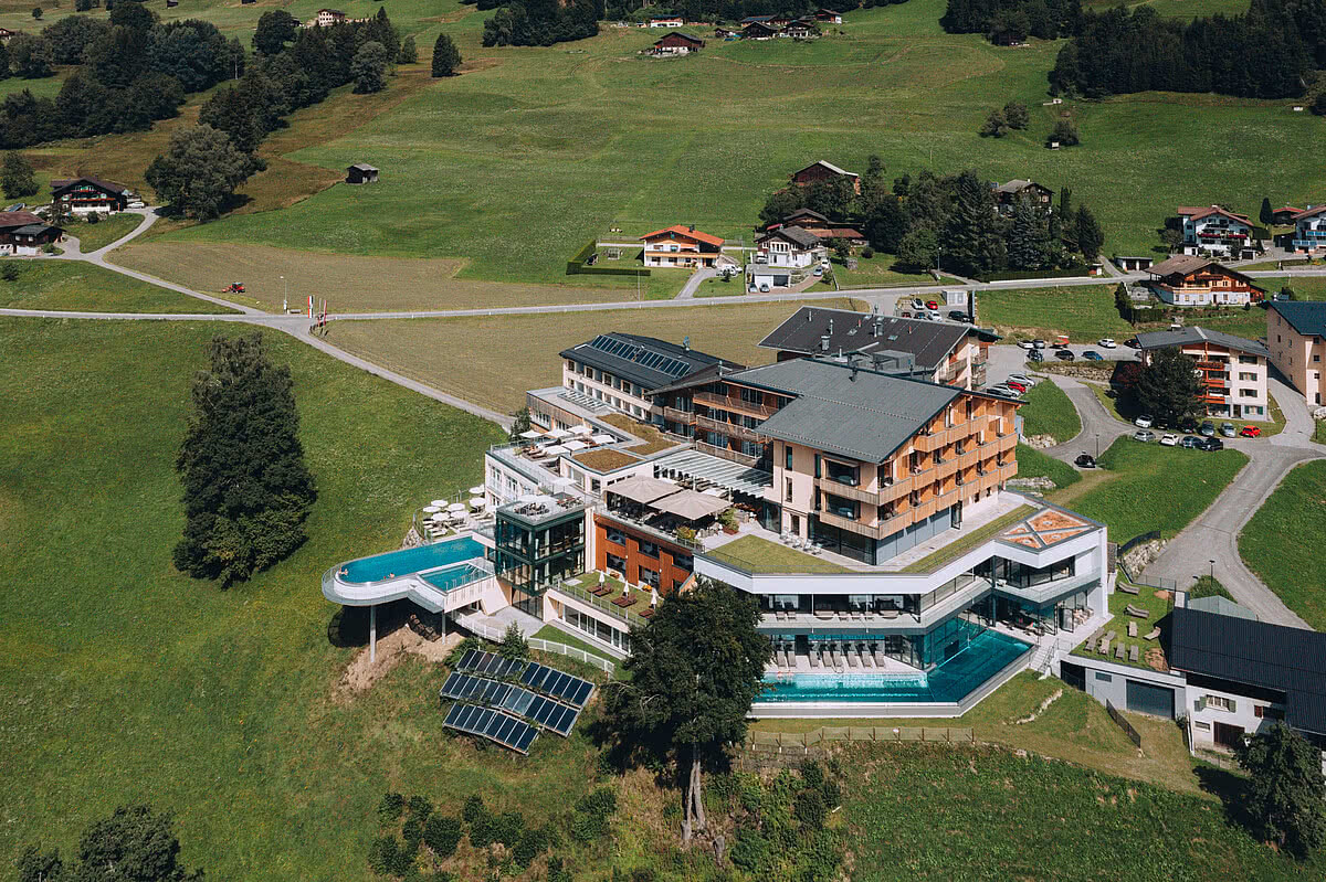 Ein großes Hotel mit Pool liegt in einer idyllischen, bergigen Landschaft. Umgeben von grünen Feldern und nahe gelegenen Gebäuden fügt es sich harmonisch in die Natur ein.