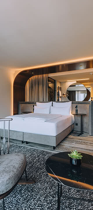 Ein elegantes Hotelzimmer mit einem großen Bett, umgeben von modernen Holzmöbeln und stimmungsvoller Beleuchtung. Eine gemütliche Sitzecke bietet Aussicht auf das stilvolle, warme Ambiente.